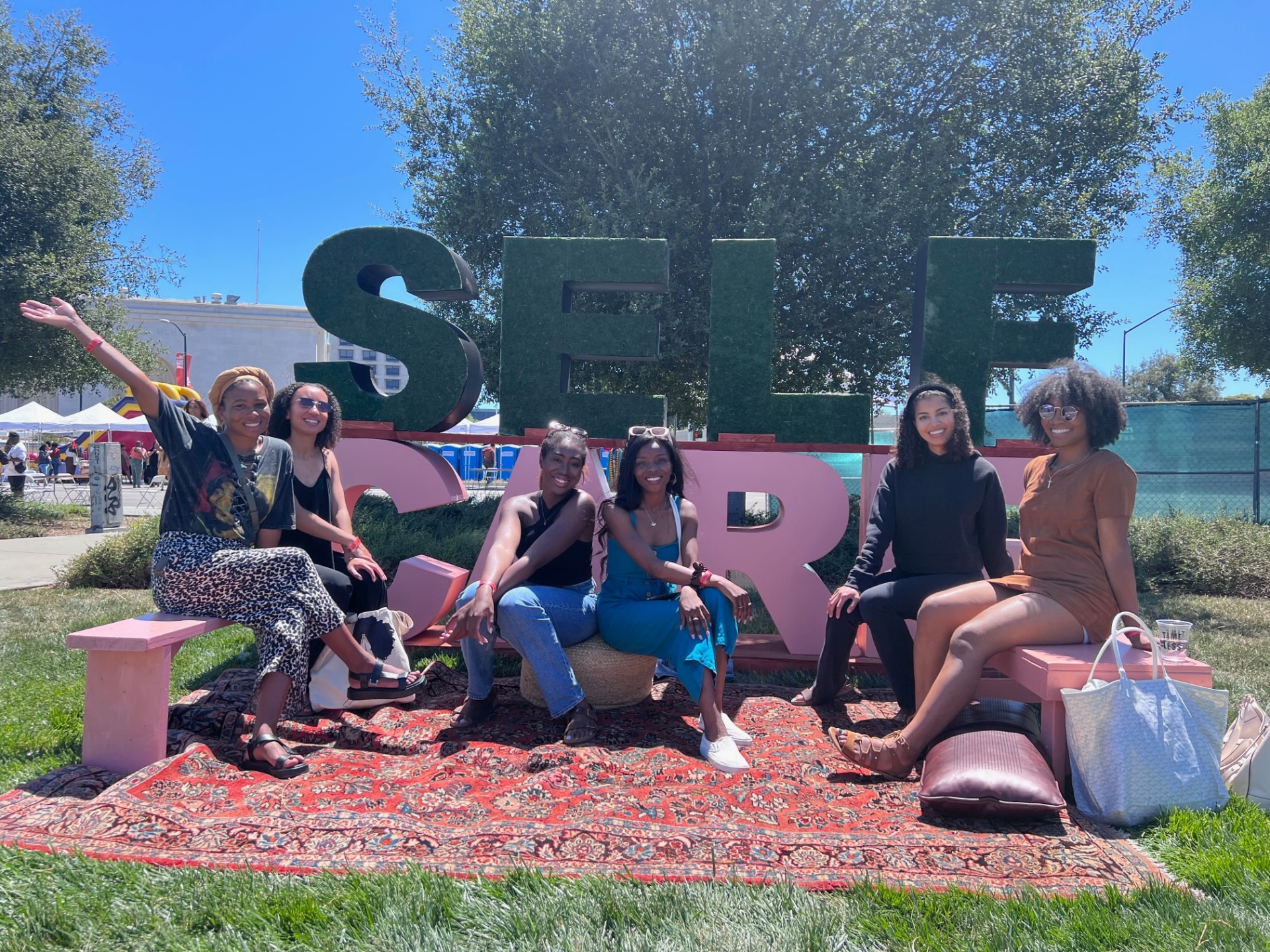 A group of Black femmes seated together at a Self Care festival in front of large mossy letters